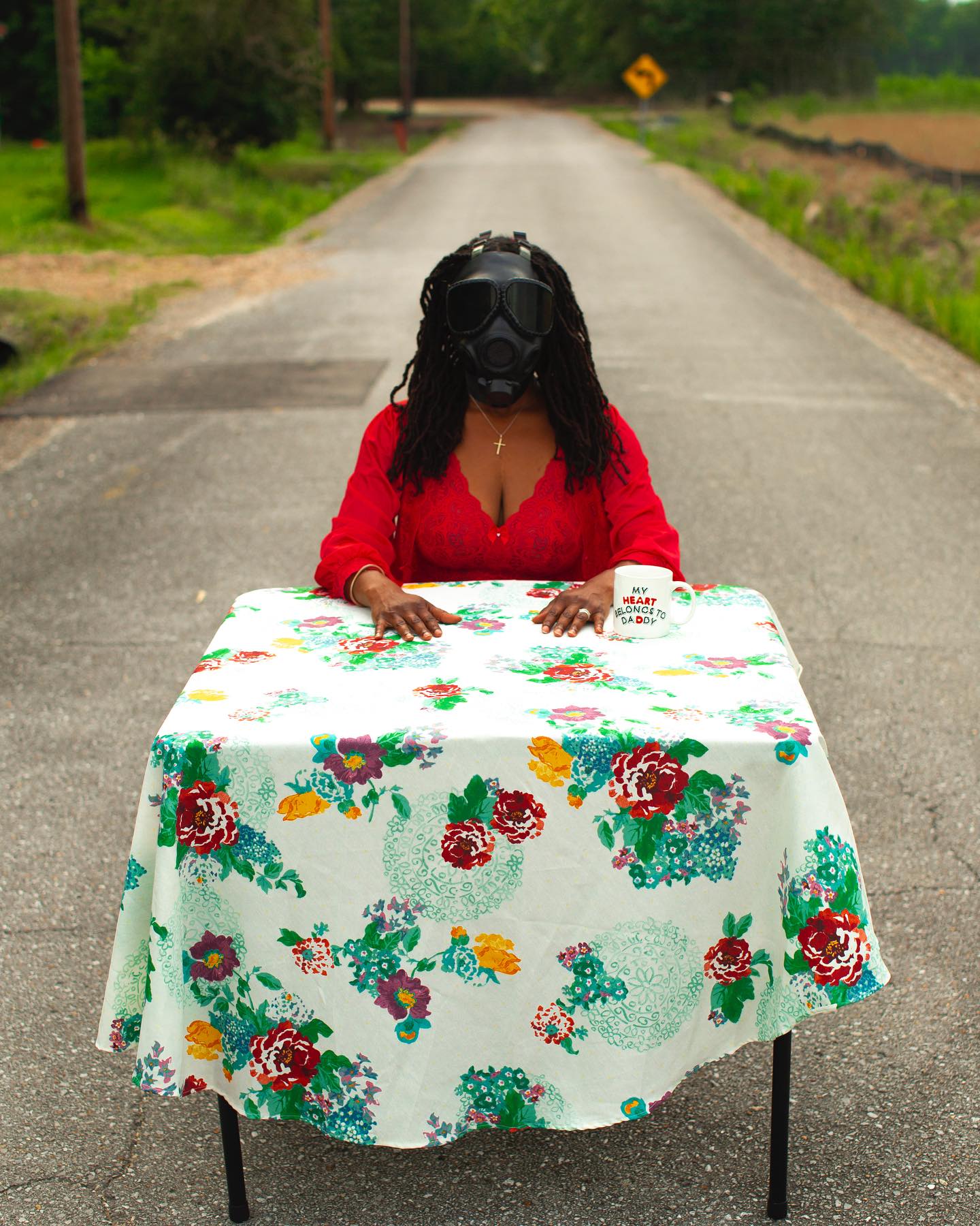 Individual seated at floral-covered table on road wearing a gas mask.