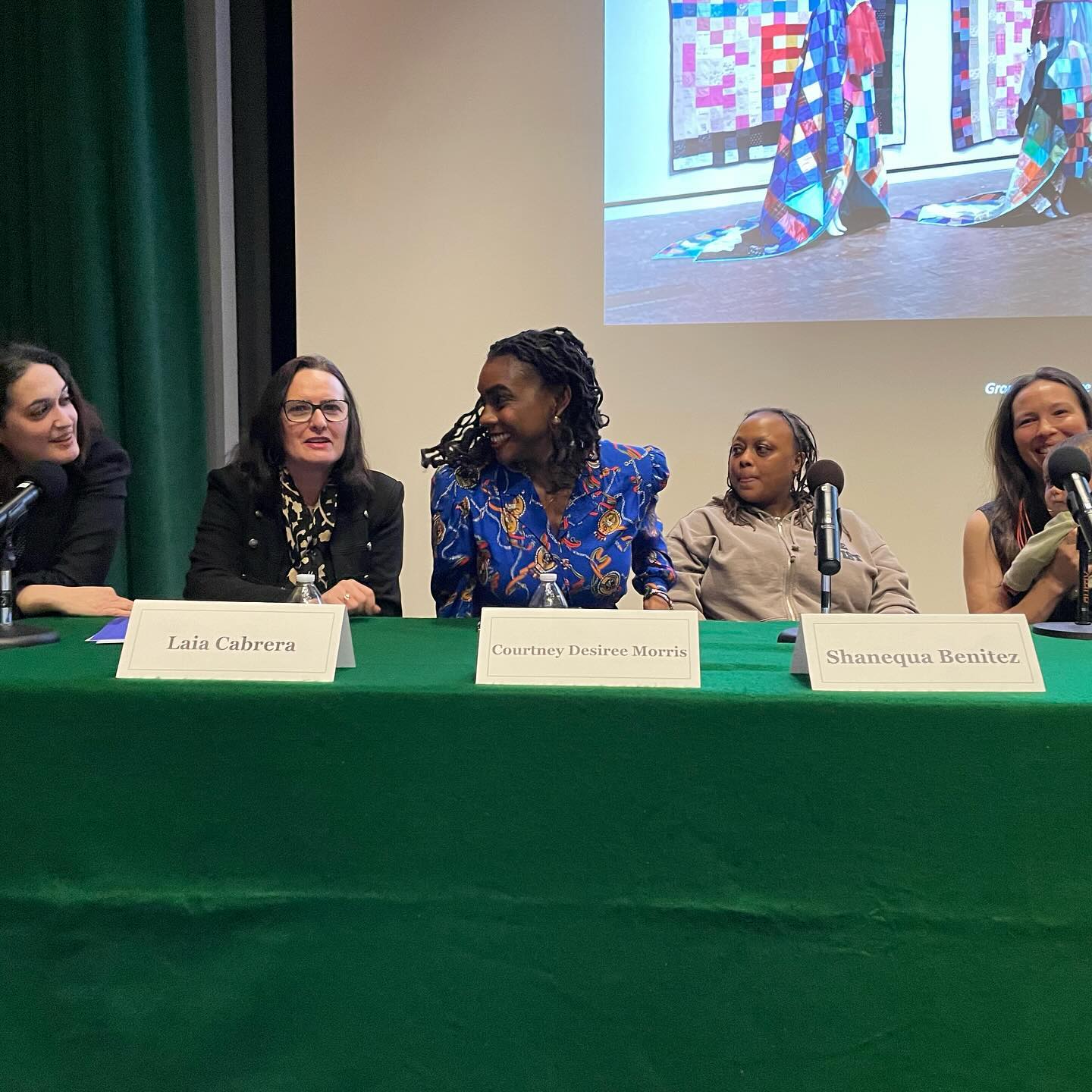 Four women are sitting at a table in front of a green screen, with one woman on the left, one in the middle, and two on the right. They are all wearing headphones and appear to be engaged in a discussion or presentation.