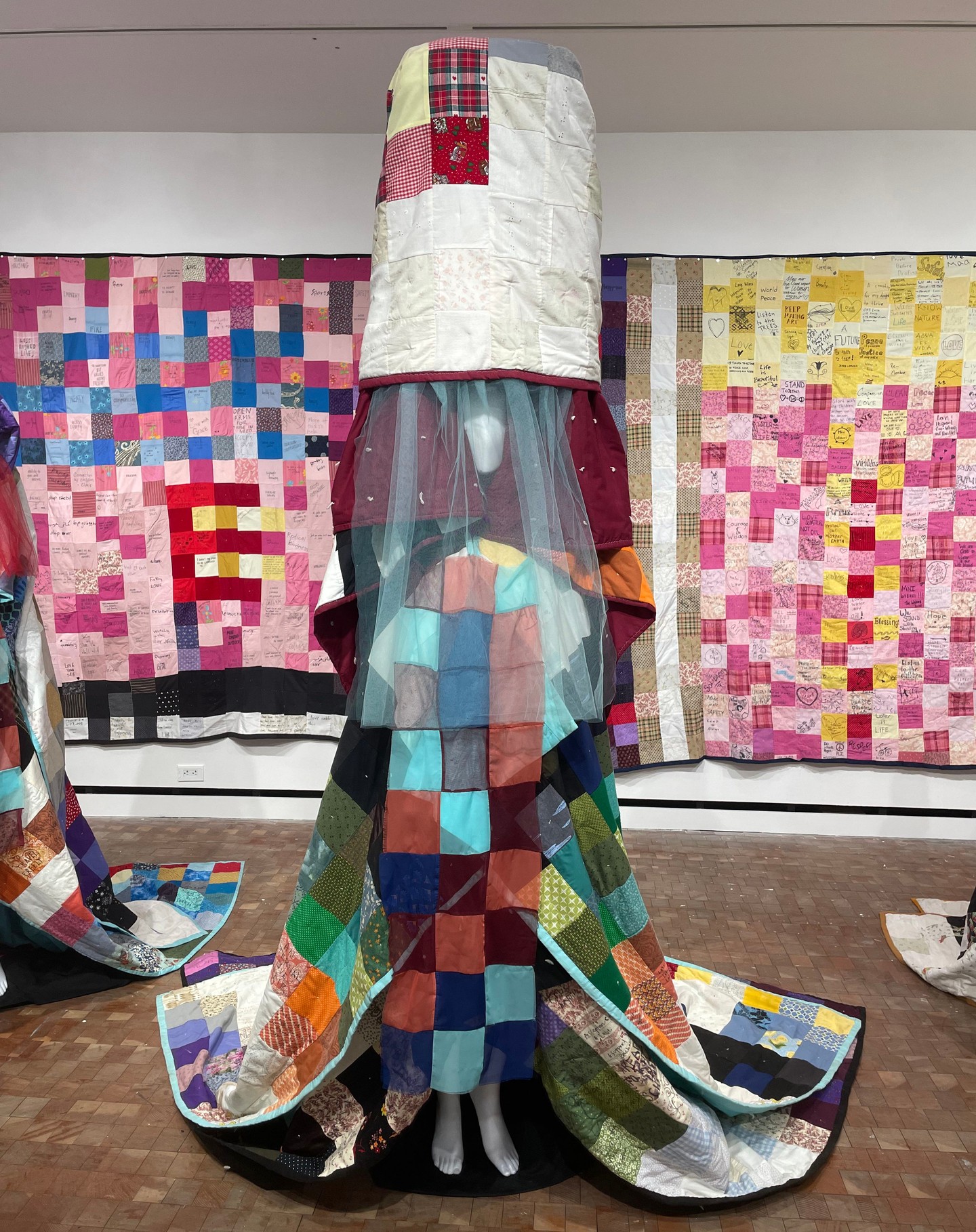 Mannequin draped in colorful patchwork quilts with a white quilt headpiece.