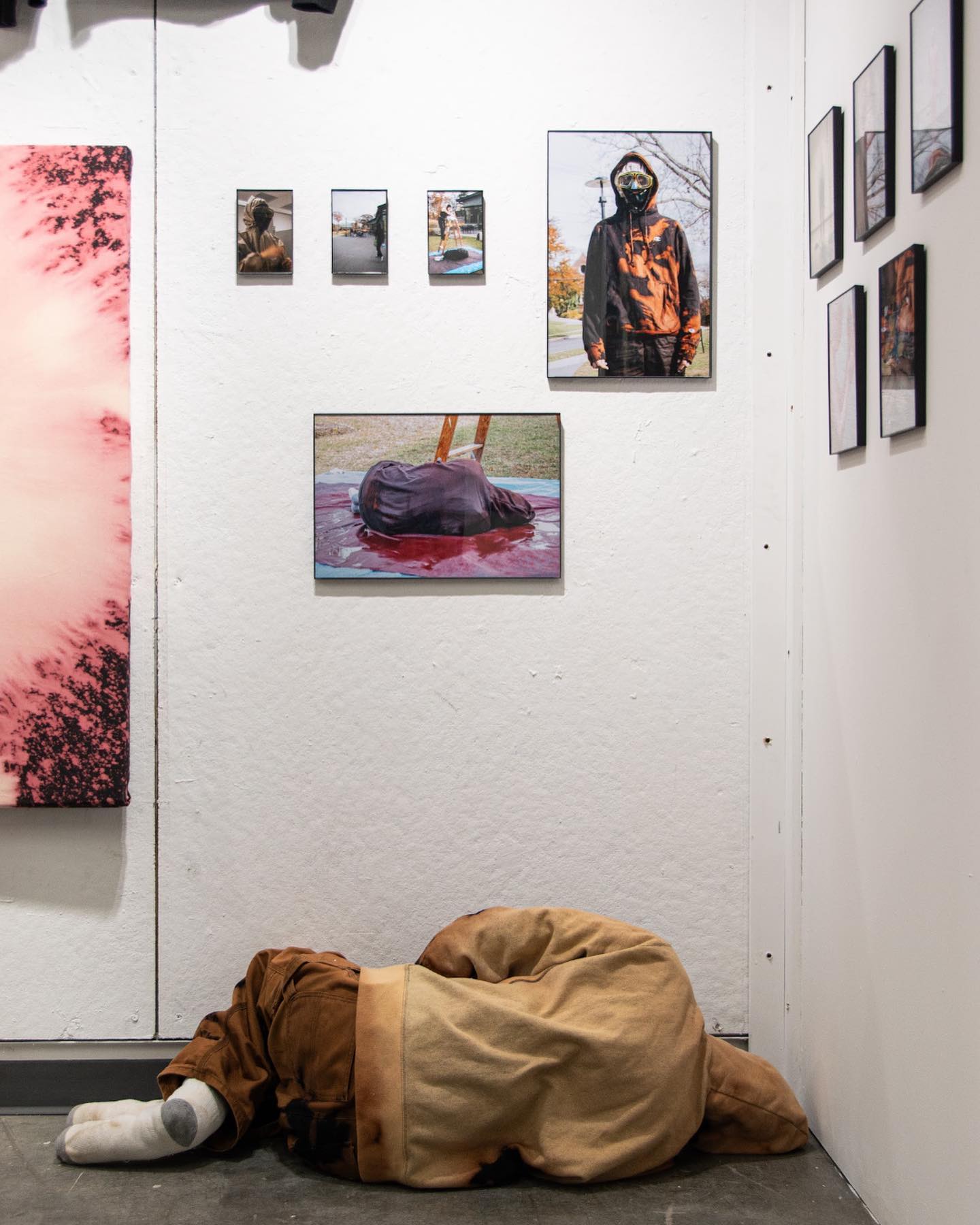 Artwork on gallery wall with a figure in a sleeping bag on the floor.