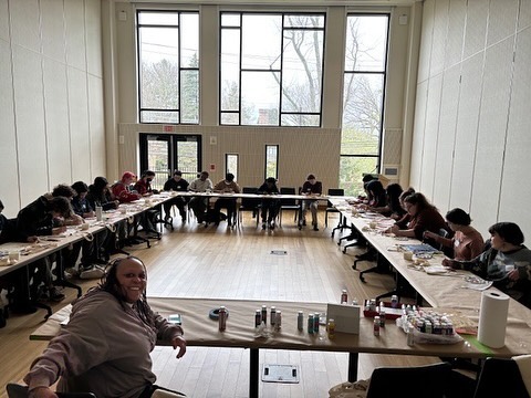 Individuals seated at long tables in a large room with big windows, some painting. One person smiling at the front.