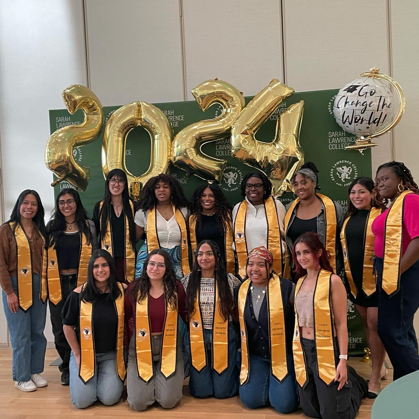 Group in graduation sashes stands before &quot;2024&quot; balloons and a &quot;Go Conquer the World&quot; sign.