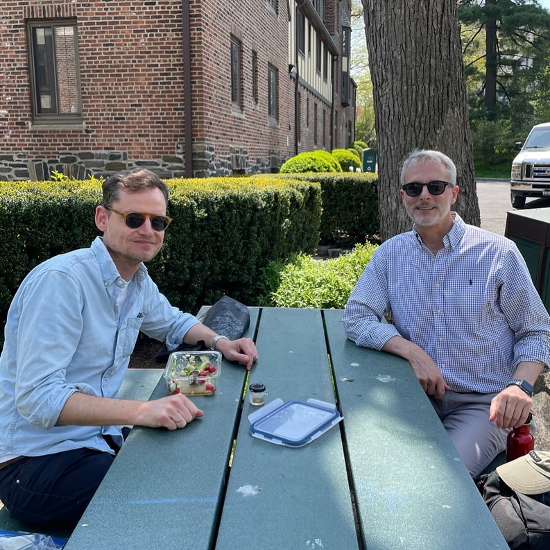 Two individuals sitting at an outdoor table with a meal, in front of a brick building.