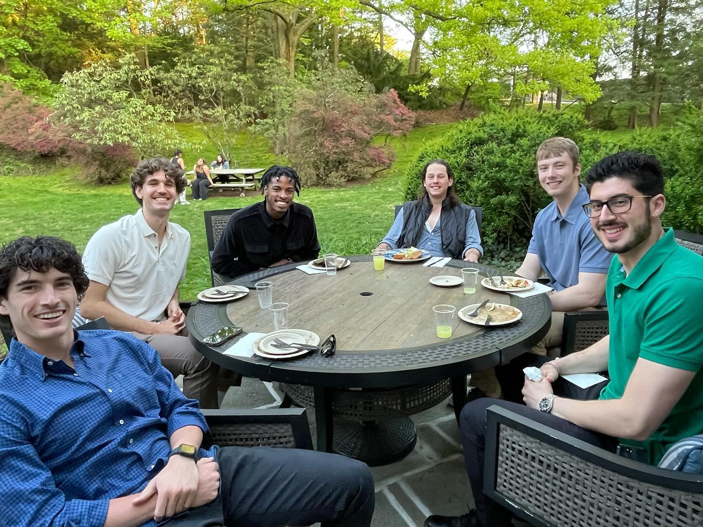 Group of people smiling at a round outdoor dining table with food and drinks.