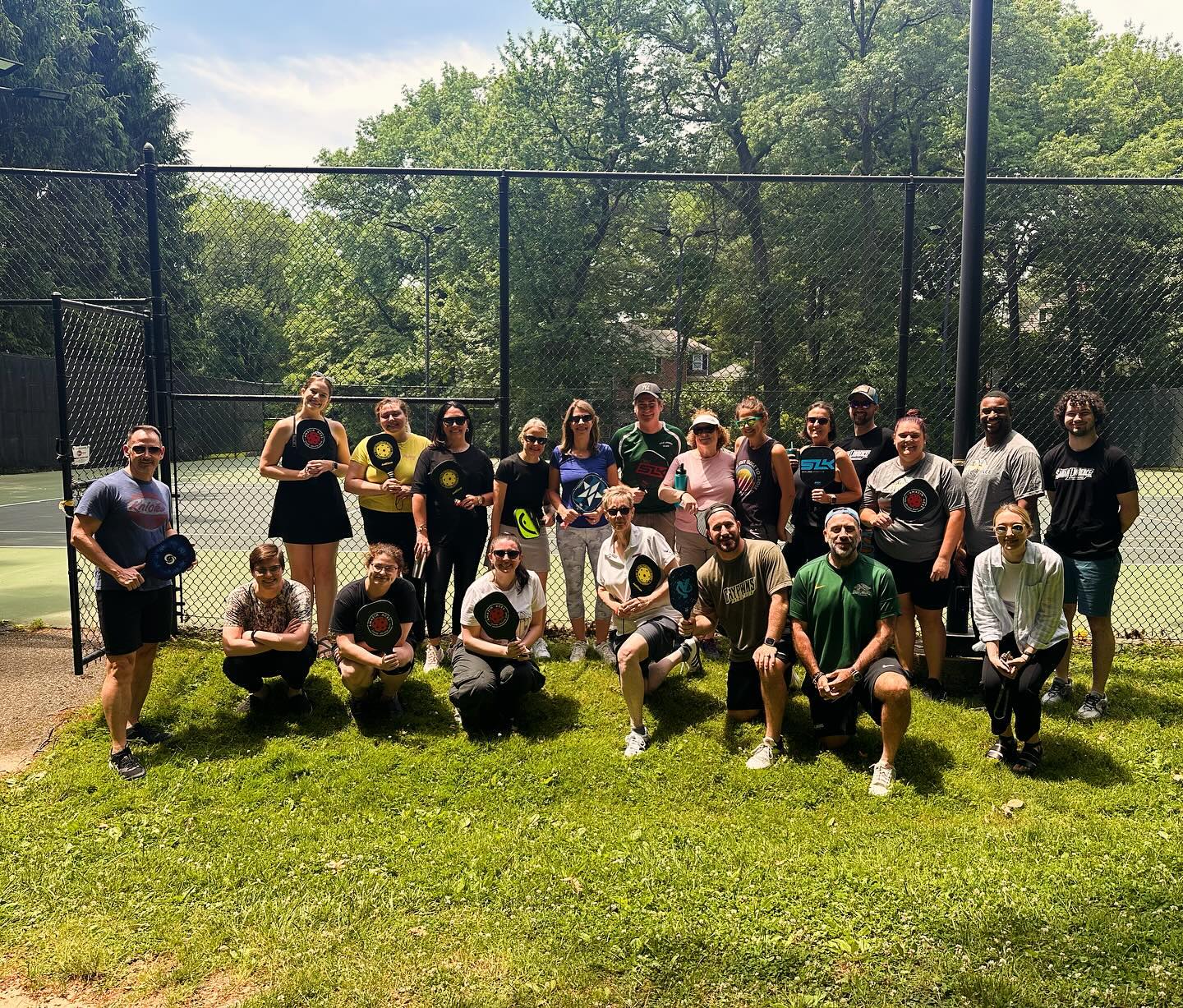 Group of people posing together with sports equipment on a sunny day at a tennis court.