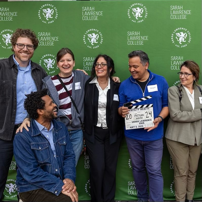 Group of people smiling at an event, one holding a clapperboard, with a logo backdrop.