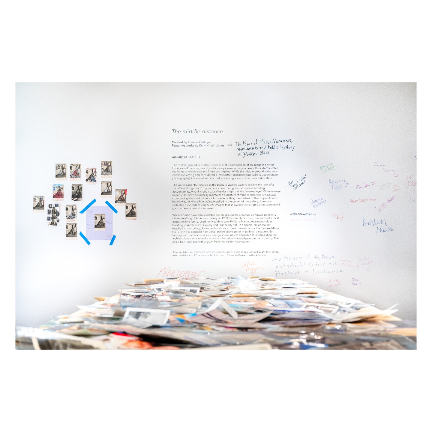 Gallery wall with photos and text. A table displays overlapping papers. Handwritten notes are scattered on the right wall.