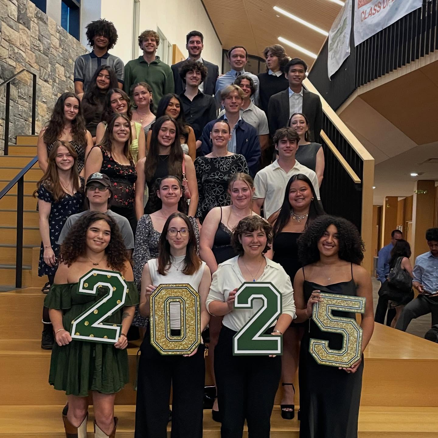 A group of people on stairs, some holding decorated numbers "2025" in an indoor setting with stone walls and wood elements.