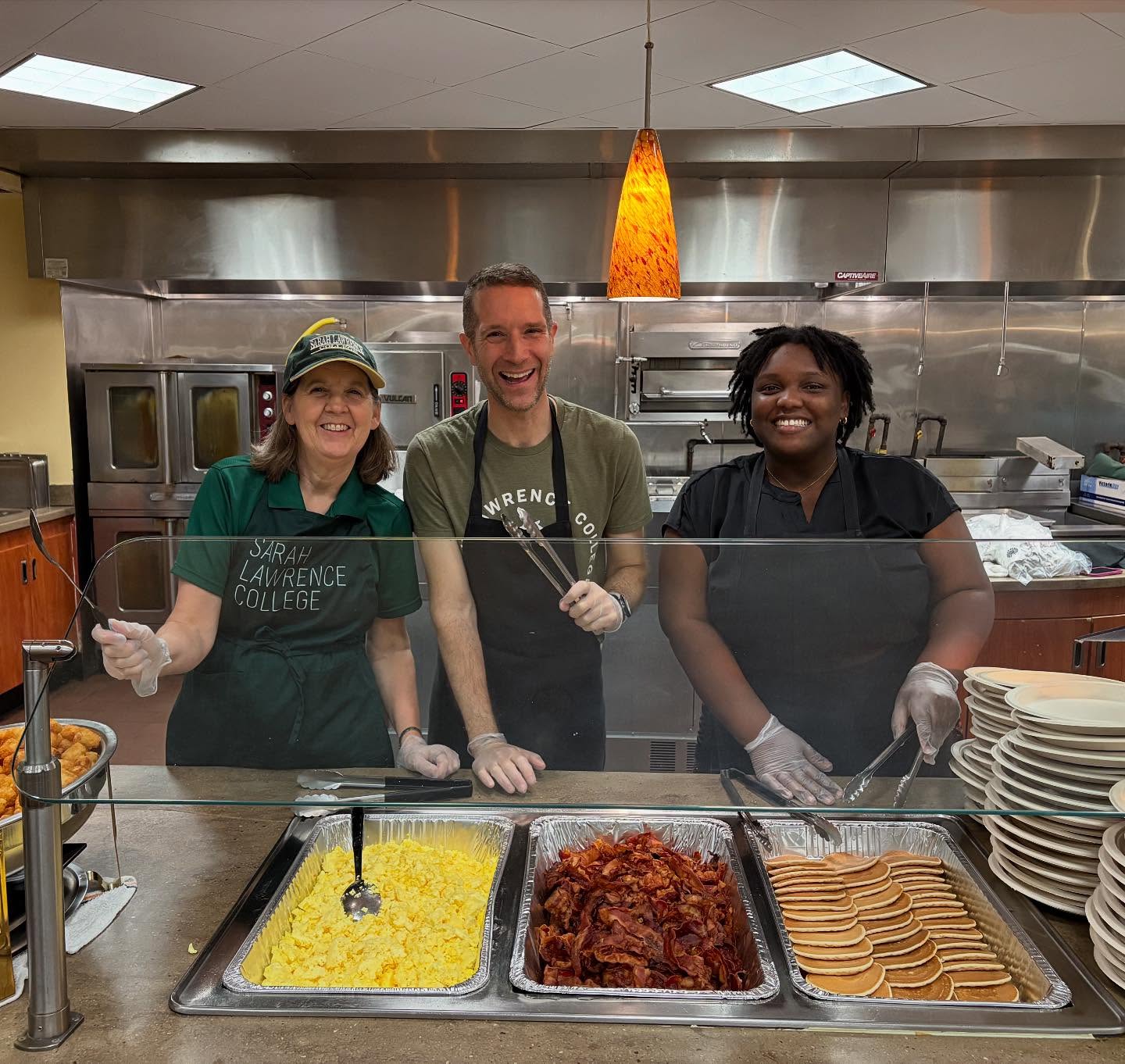Three people smiling behind a cafeteria counter with trays of scrambled eggs, bacon, and pancakes. Plates are stacked on the side.
