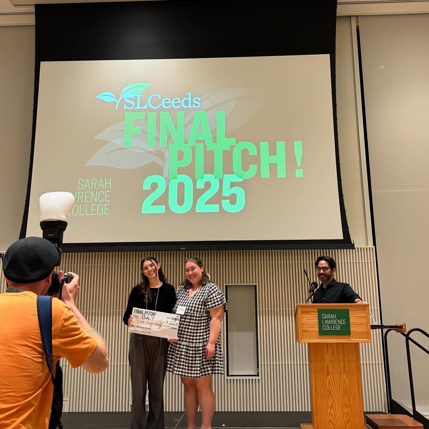Two individuals smiling on stage, holding an oversized check under a large "SLCeeds Final Pitch 2025" screen while a photographer captures the moment.