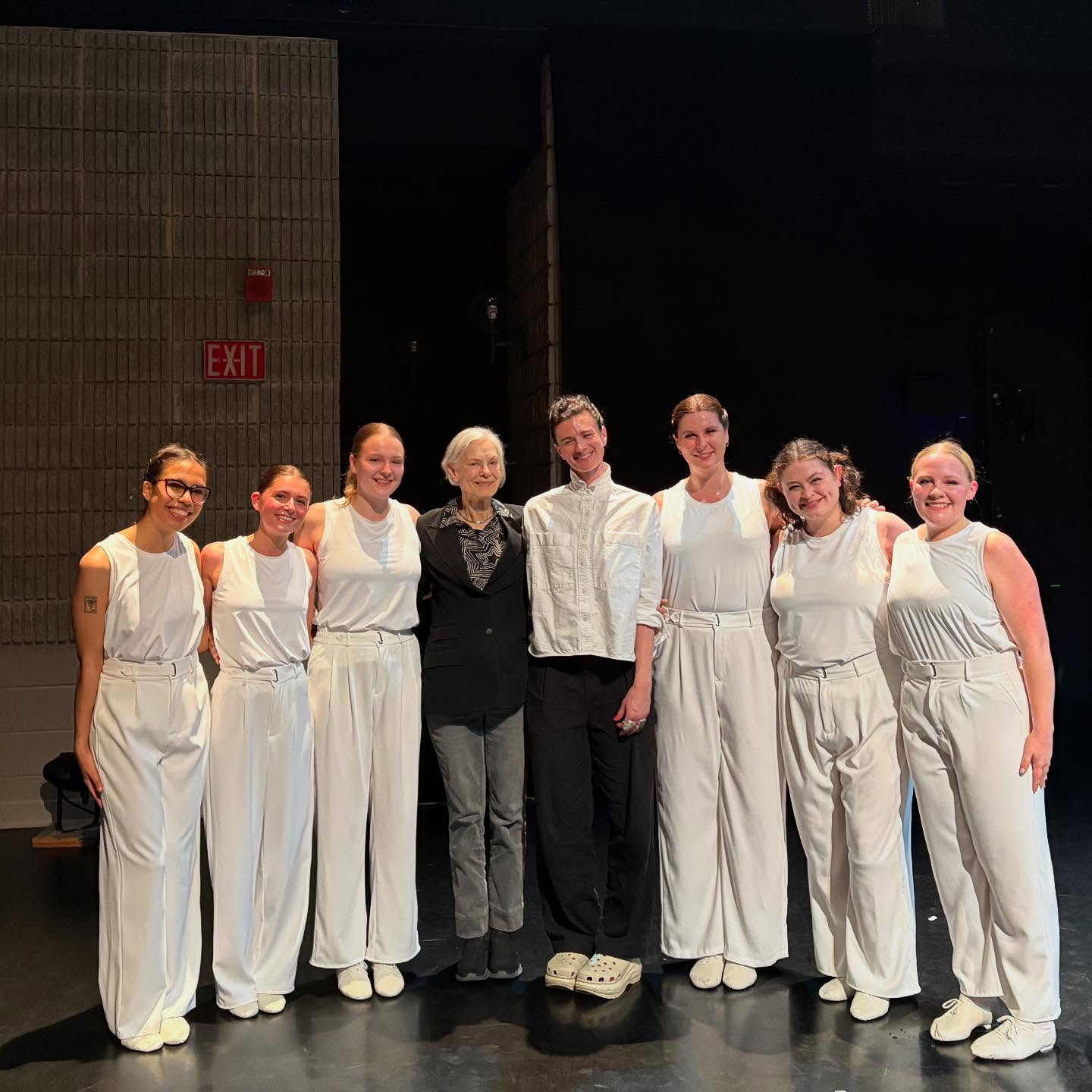 A group of eight people pose on a dimly lit stage. Seven wear white outfits, and one in the center wears a dark suit.