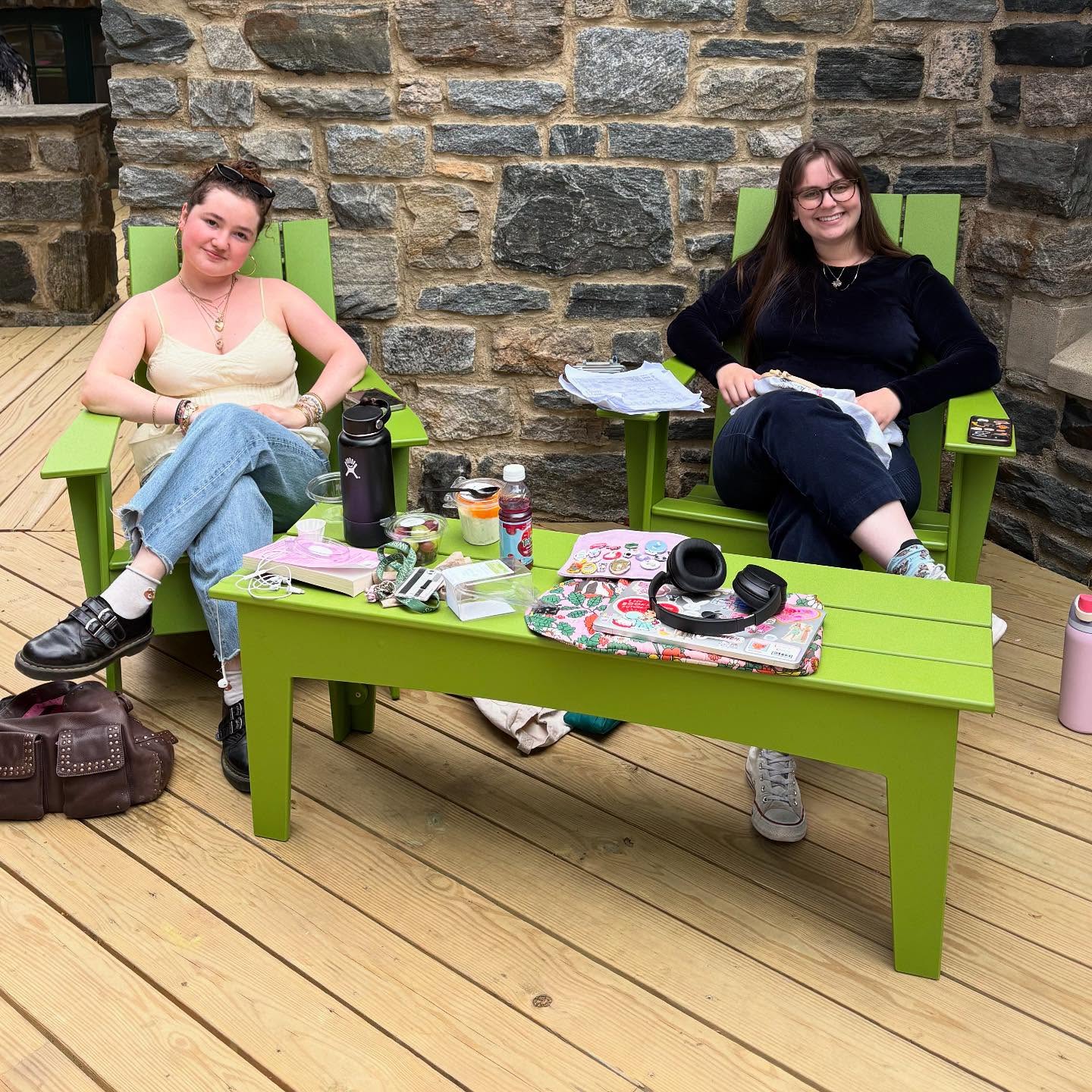 Two people sit in green chairs on a wood deck, surrounded by various items on a matching green table, with a stone wall in the background.