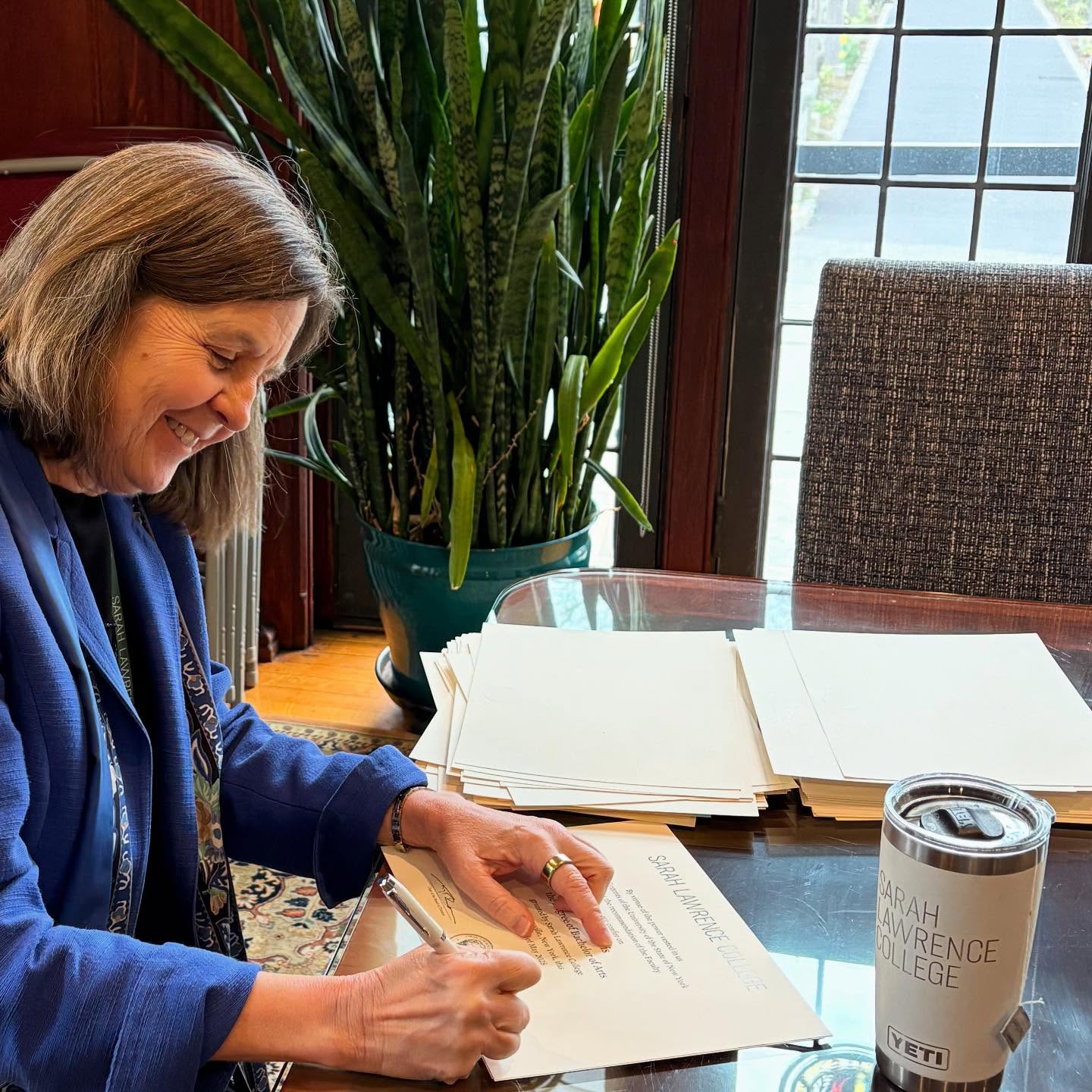 Person in a blue jacket signs papers at a table, smiling. A large plant and a travel mug marked "Sarah Lawrence College" are visible nearby.