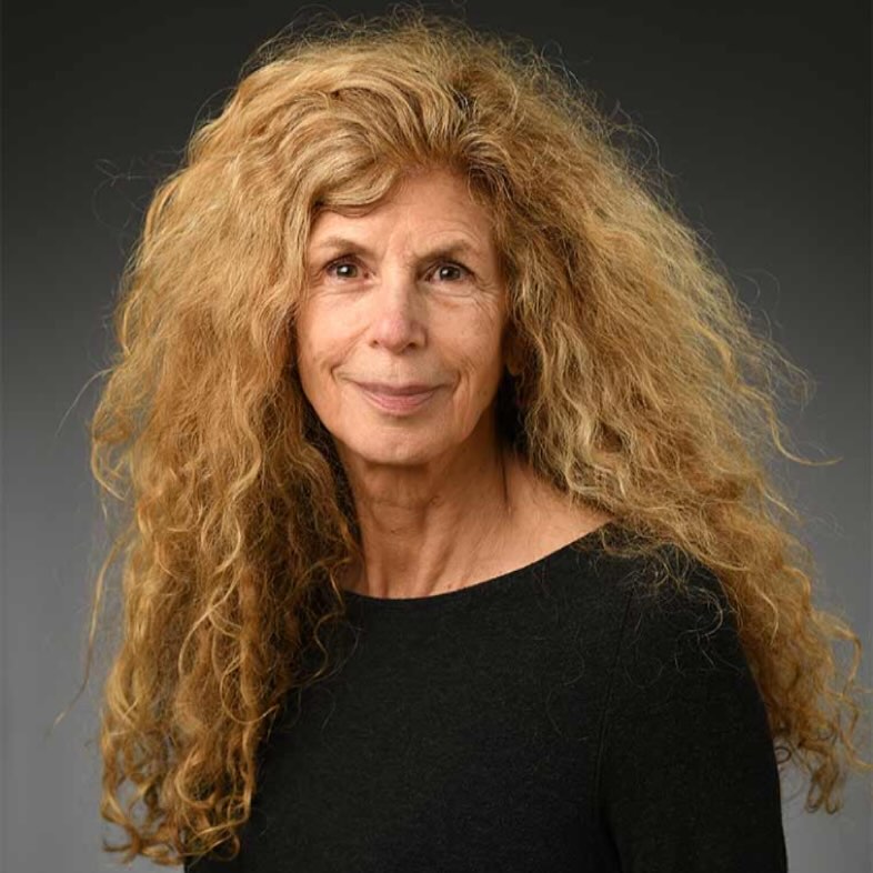 Individual with long, curly hair and a neutral expression, wearing a dark top, against a gradient gray background.
