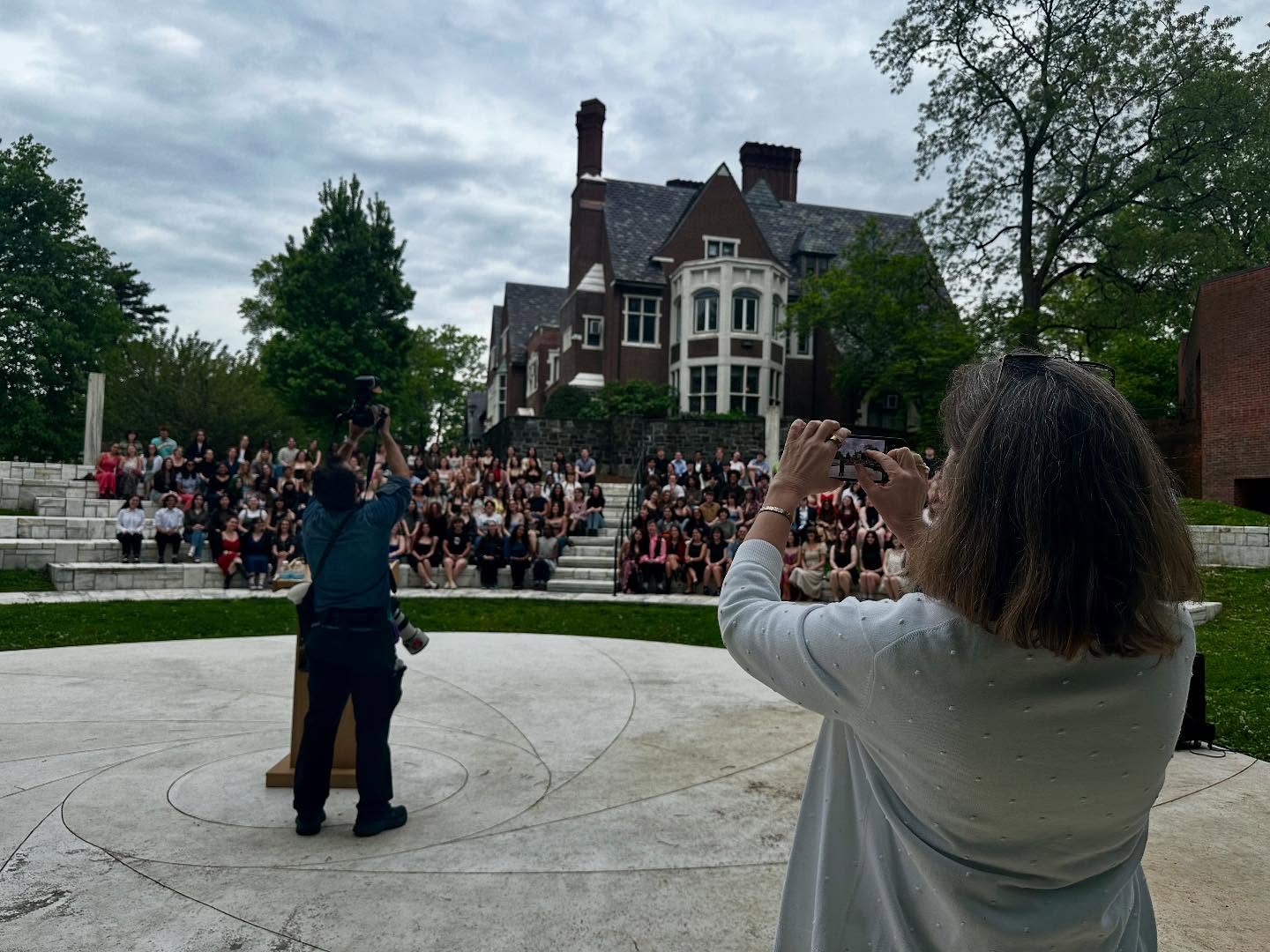 A person photographs a large group seated on tiered steps, in front of a large historic building, surrounded by trees.