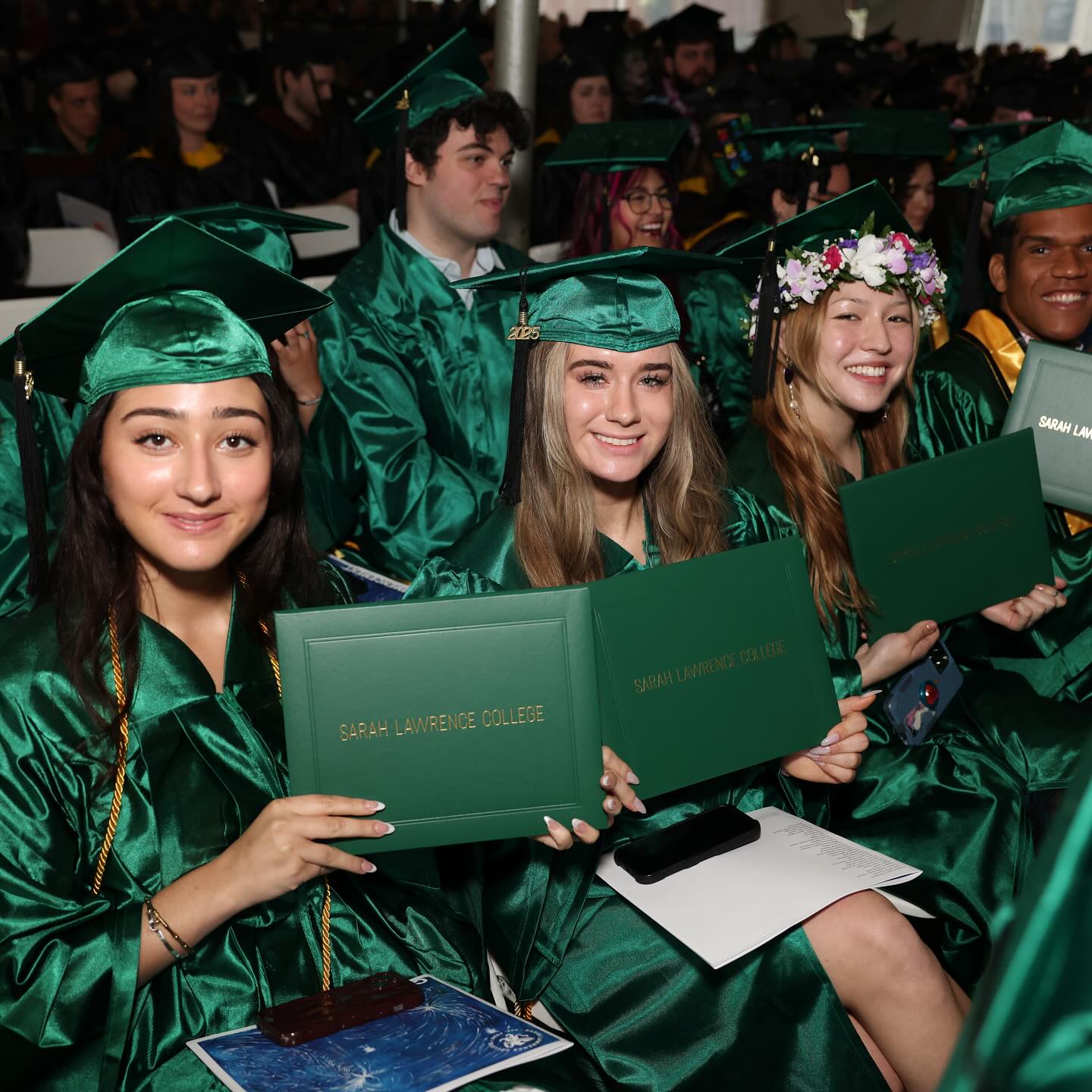 Graduates in green gowns and caps hold diplomas at a Sarah Lawrence College ceremony, smiling and sitting closely together.