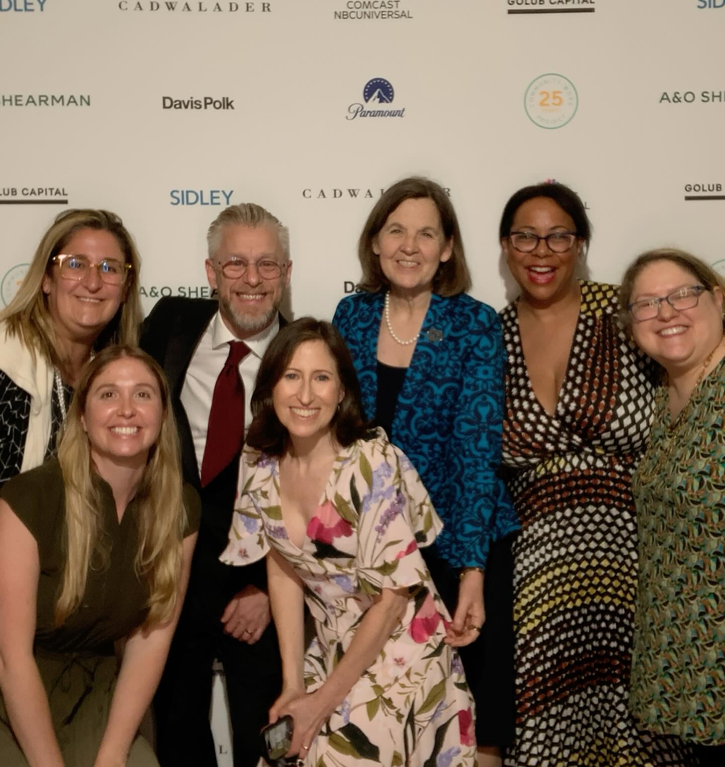 A group of seven people smiling at a social event, standing in front of a white backdrop with various logos.