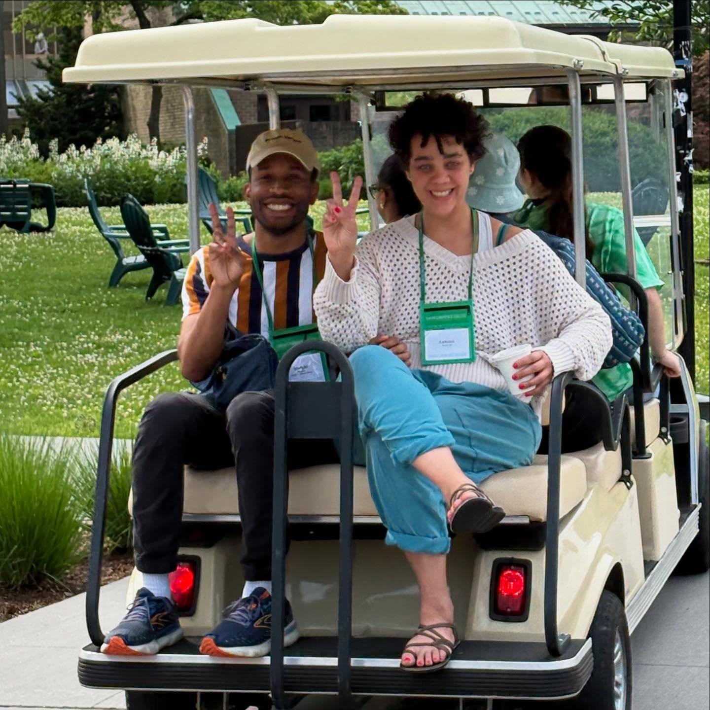 Two people sitting on the back of a golf cart, smiling and making peace signs, with greenery in the background.
