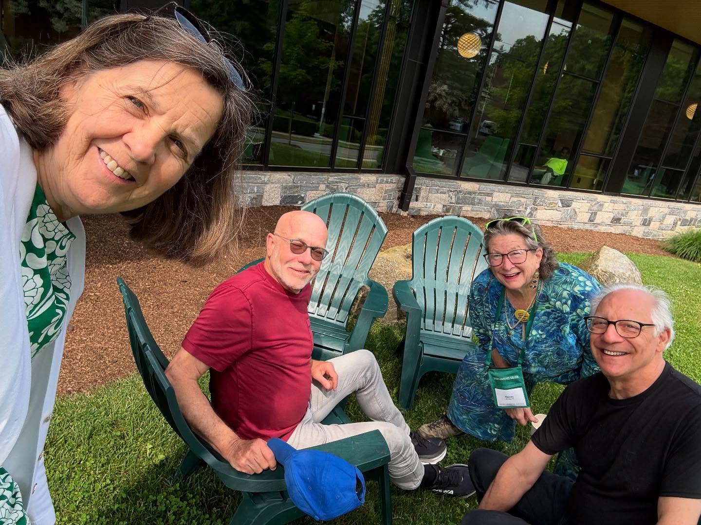 Four people smiling together, seated outdoors on green chairs near a building with large windows.