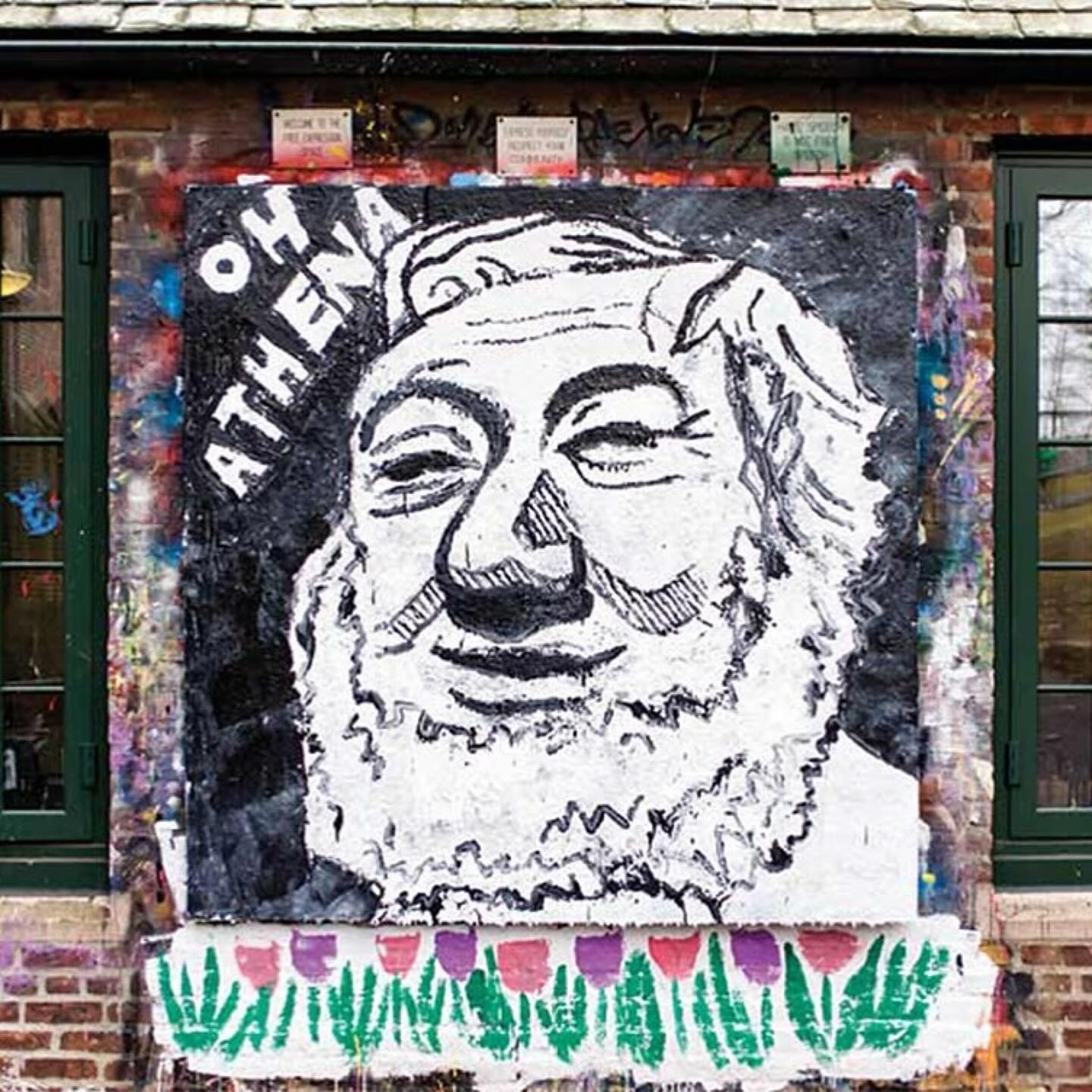 Graffiti on a brick wall depicts a face with a beard and the text "OH ATHENA." Below, painted flowers are visible.