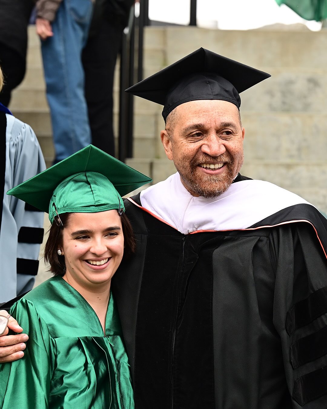 Two people in graduation attire, one in green robes and cap, the other in black robes with a cap, smiling and standing together.