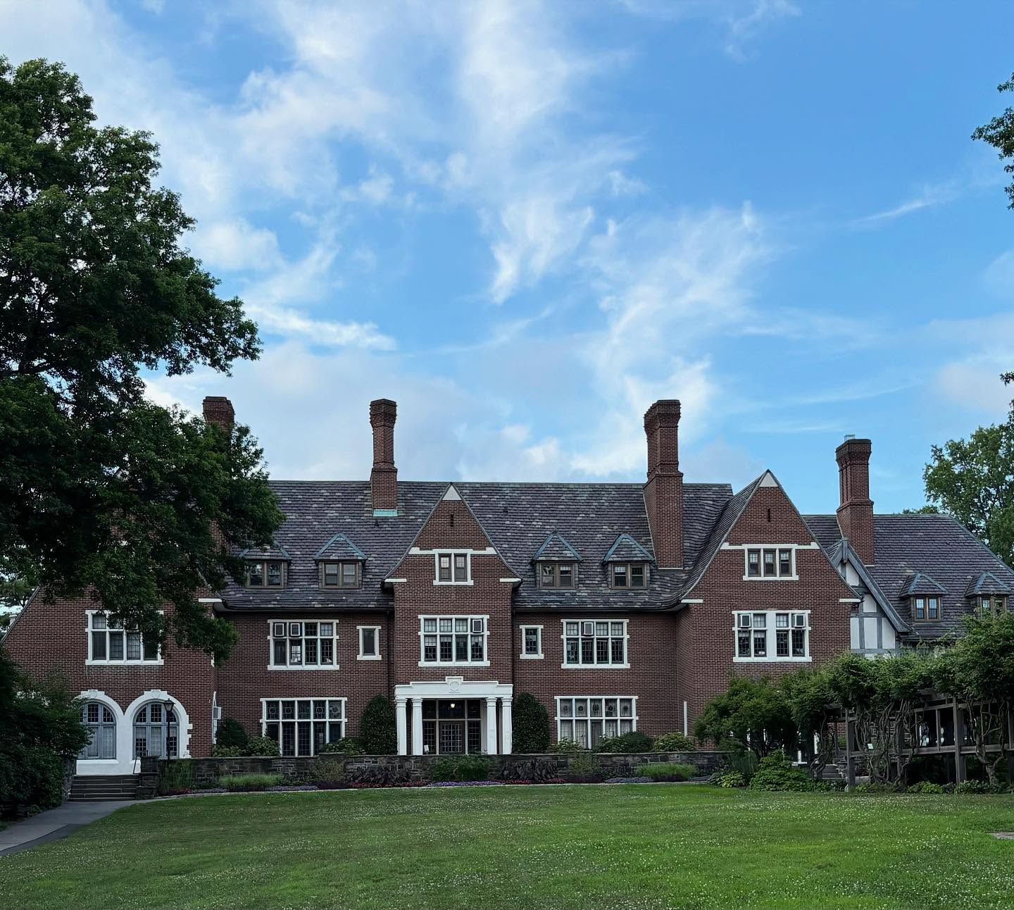 Large brick mansion with multiple chimneys, surrounded by greenery and under a partly cloudy blue sky.