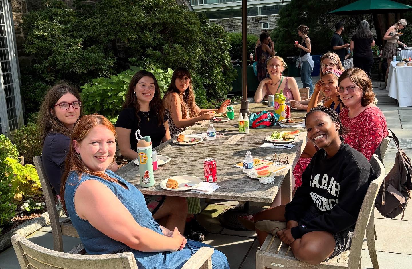A group of people sitting around a table outdoors, smiling, with food and drinks in front of them. Lush greenery and other people in the background.