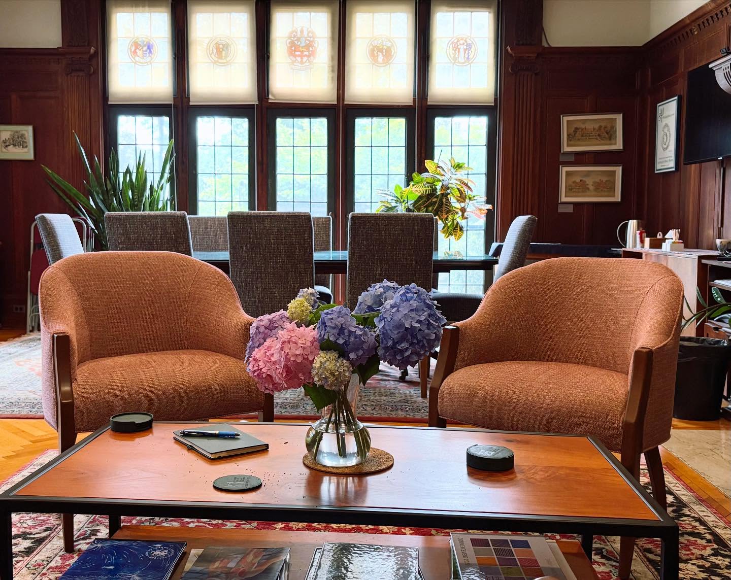 Cozy room with two orange chairs, a wooden table with hydrangeas, and large windows. Plants and framed art adorn the background.