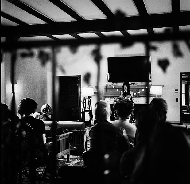 Person speaking to an audience in a dimly lit room with a TV and lamps on the wall; people sit facing the speaker.