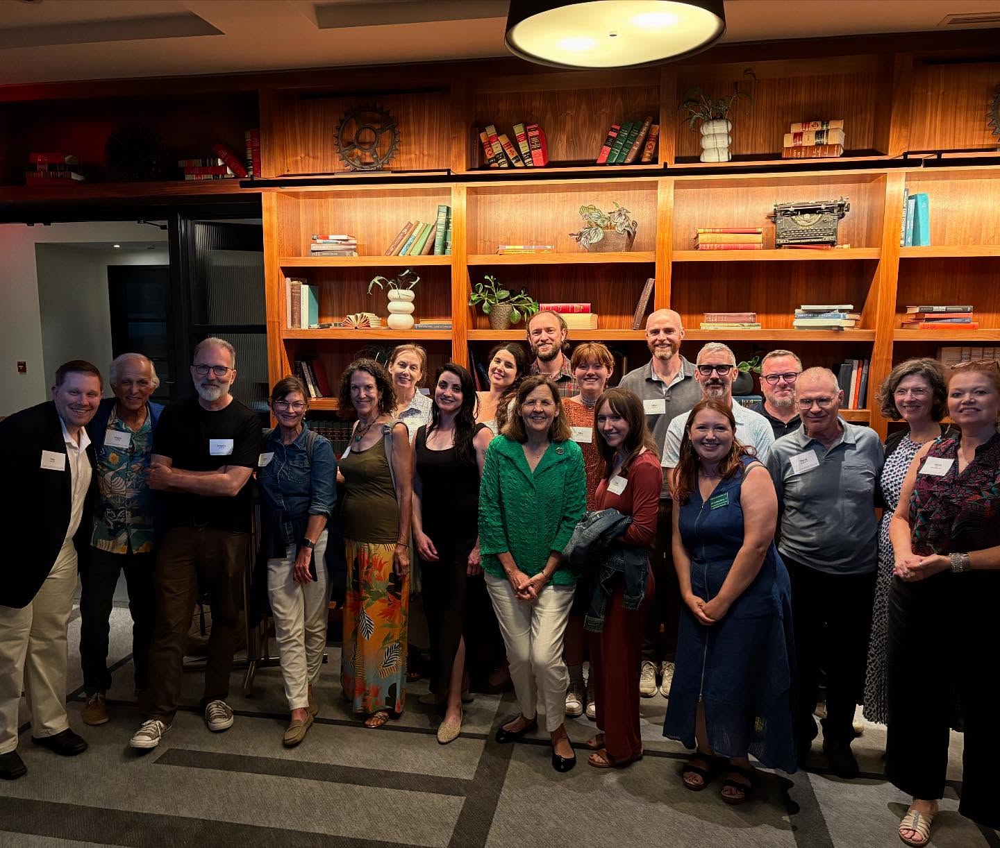 A group of people smiling in front of a wooden bookshelf with books and plants, in a warmly lit room.
