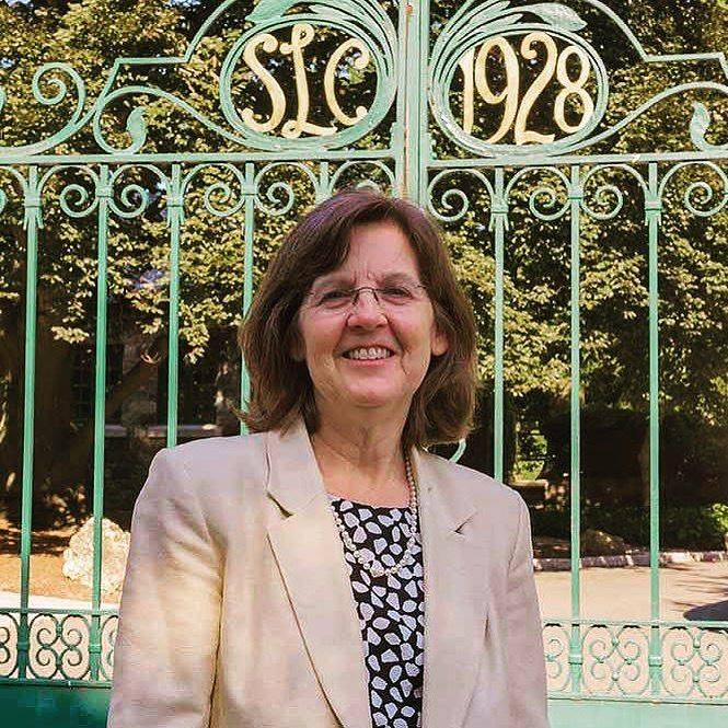 Person in a light blazer stands smiling in front of a green gate with "SLC 1928" inscription. Trees are visible in the background.