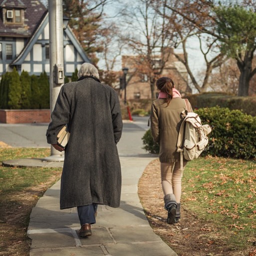 Two people walking on a sidewalk, one carrying books and the other a backpack. Trees and houses line the street in the background.