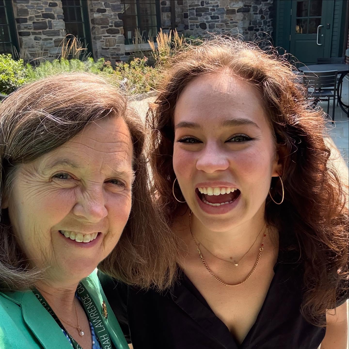 Two people smiling outdoors in front of a stone building and greenery.