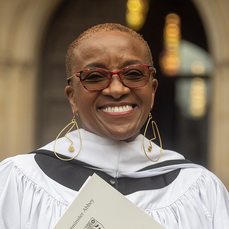 A person wearing glasses, large earrings, and a white robe holds a booklet. They are smiling in front of a blurred background.