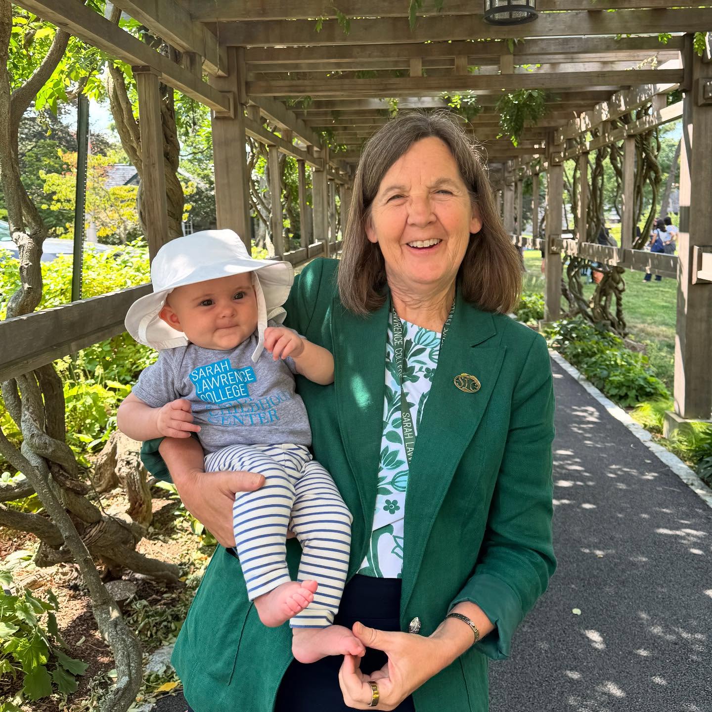 An adult in a green blazer holds a baby wearing a sun hat and striped pants, standing under a wooden trellis surrounded by greenery.