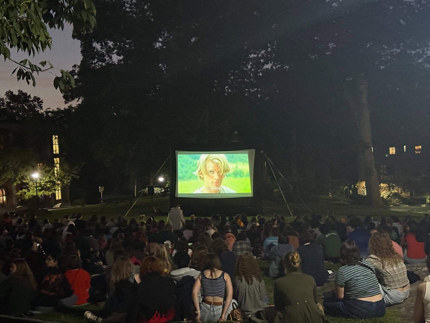 Outdoor movie screening at night. A crowd sits on the grass watching a film on a large screen under tree-lined surroundings.