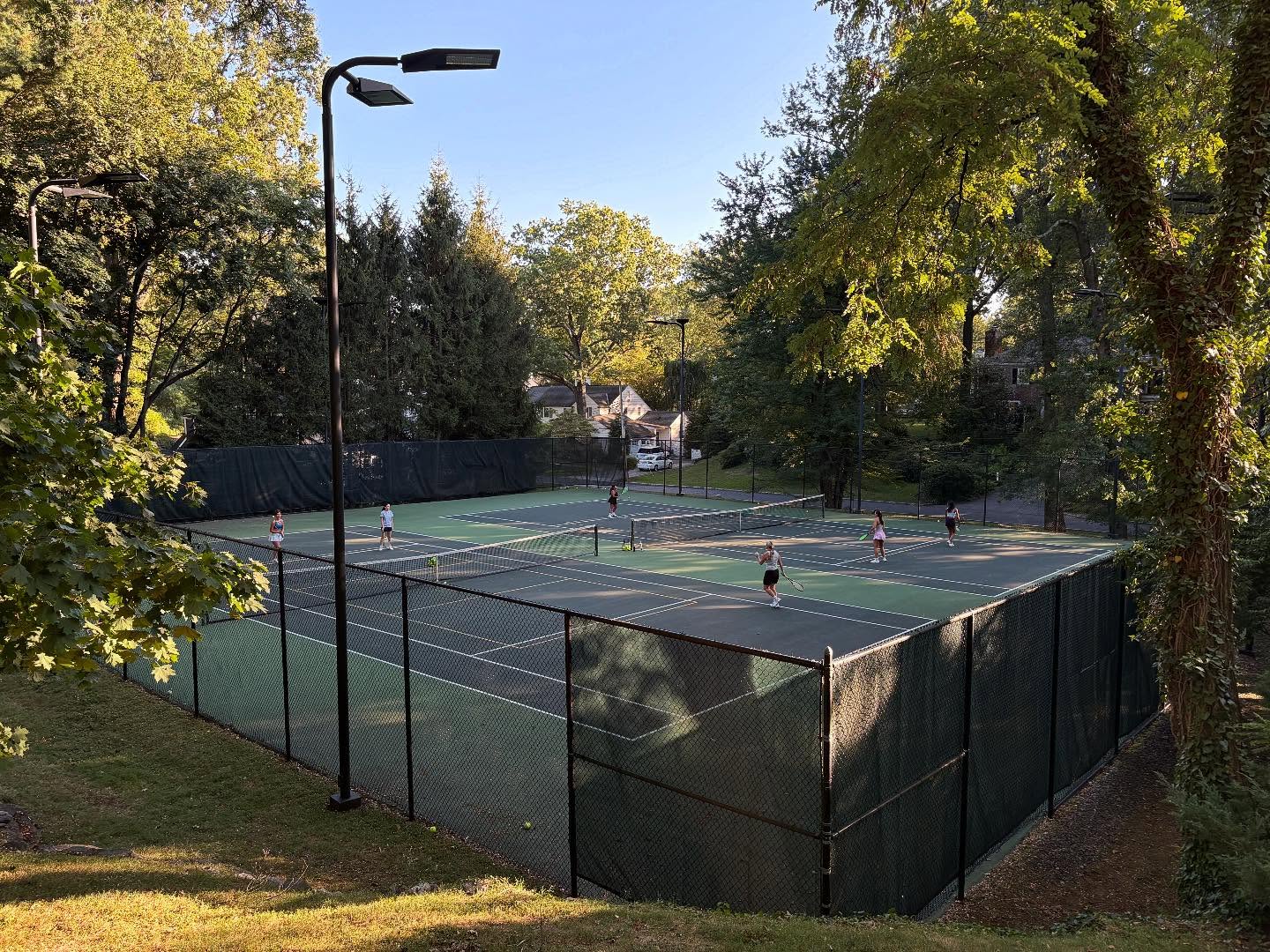 Four people are playing tennis on an outdoor court surrounded by trees, with sunlight filtering through the leaves.