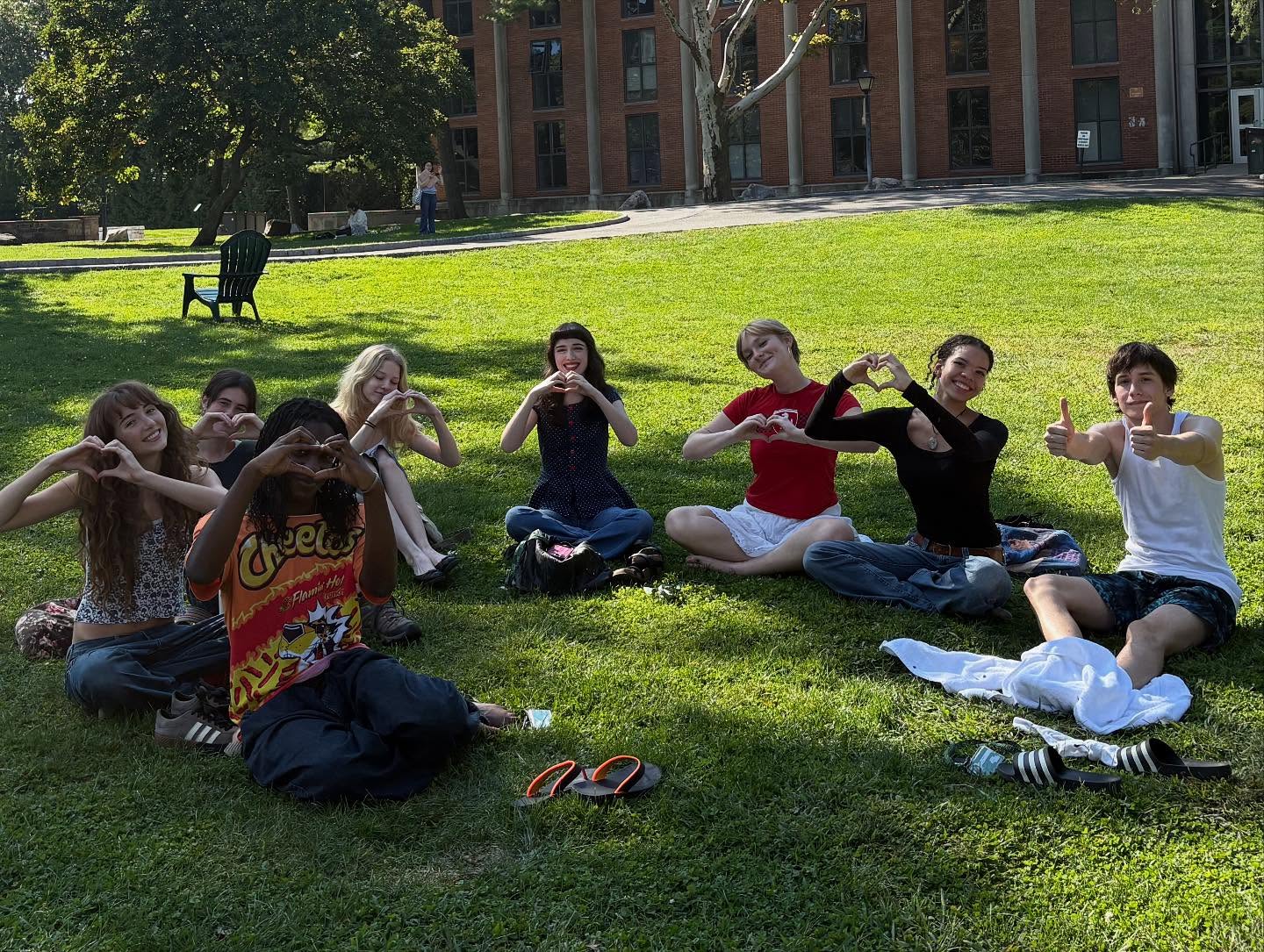Group of people sitting on grass forming heart shapes with hands and giving thumbs up. Trees and a building in the background.