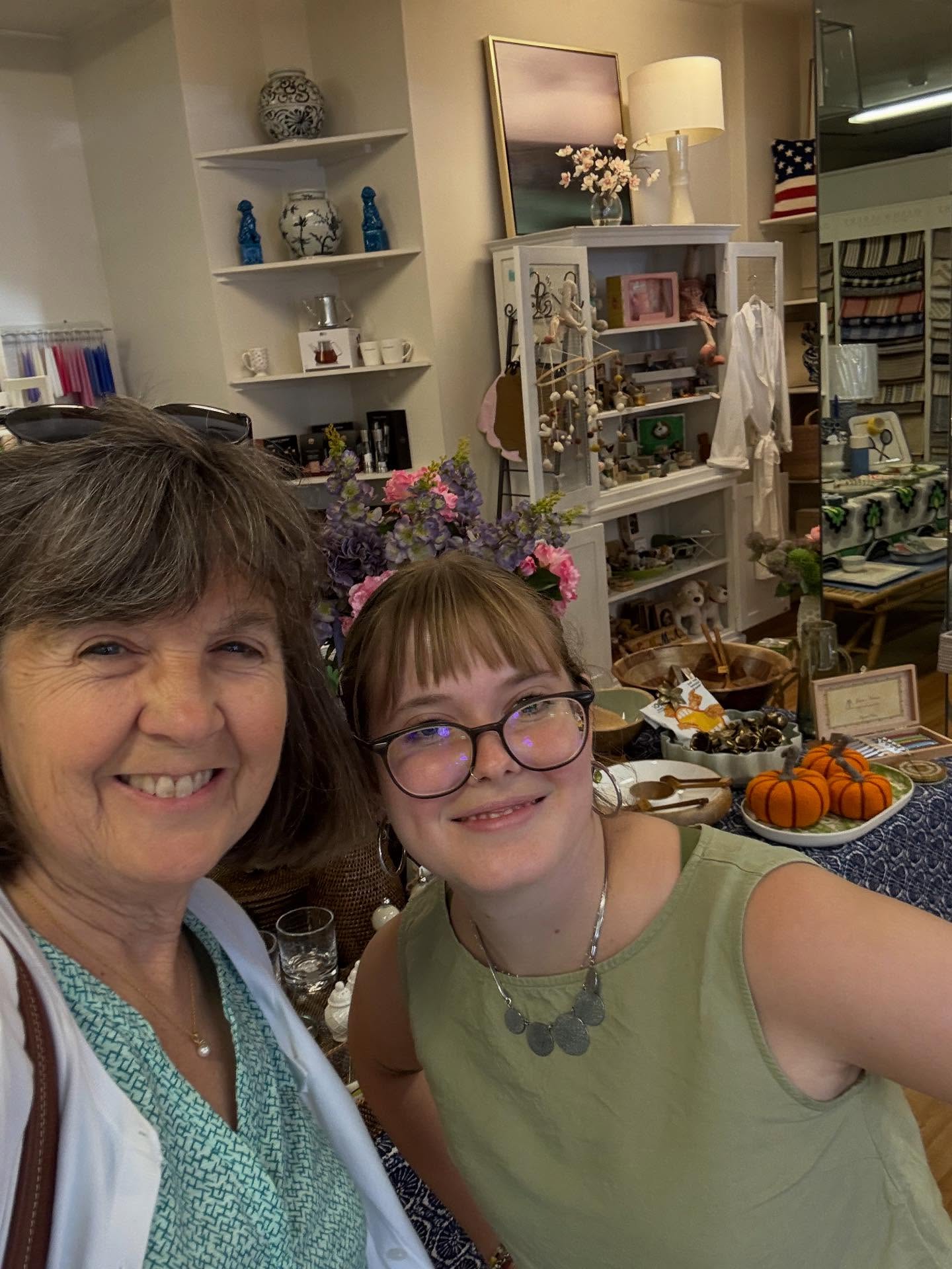 Two people smiling in a shop with shelves of decor items, art, and a table display featuring small pumpkins and flowers.