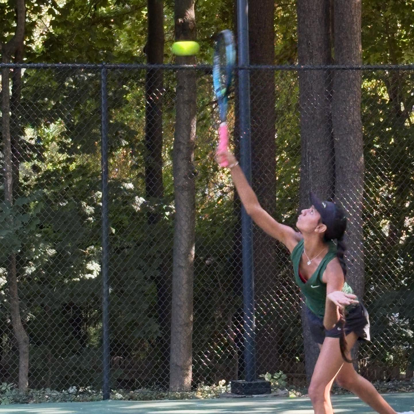 Person playing tennis on an outdoor court, preparing to hit the ball with a racquet. Trees and a chain-link fence in the background.