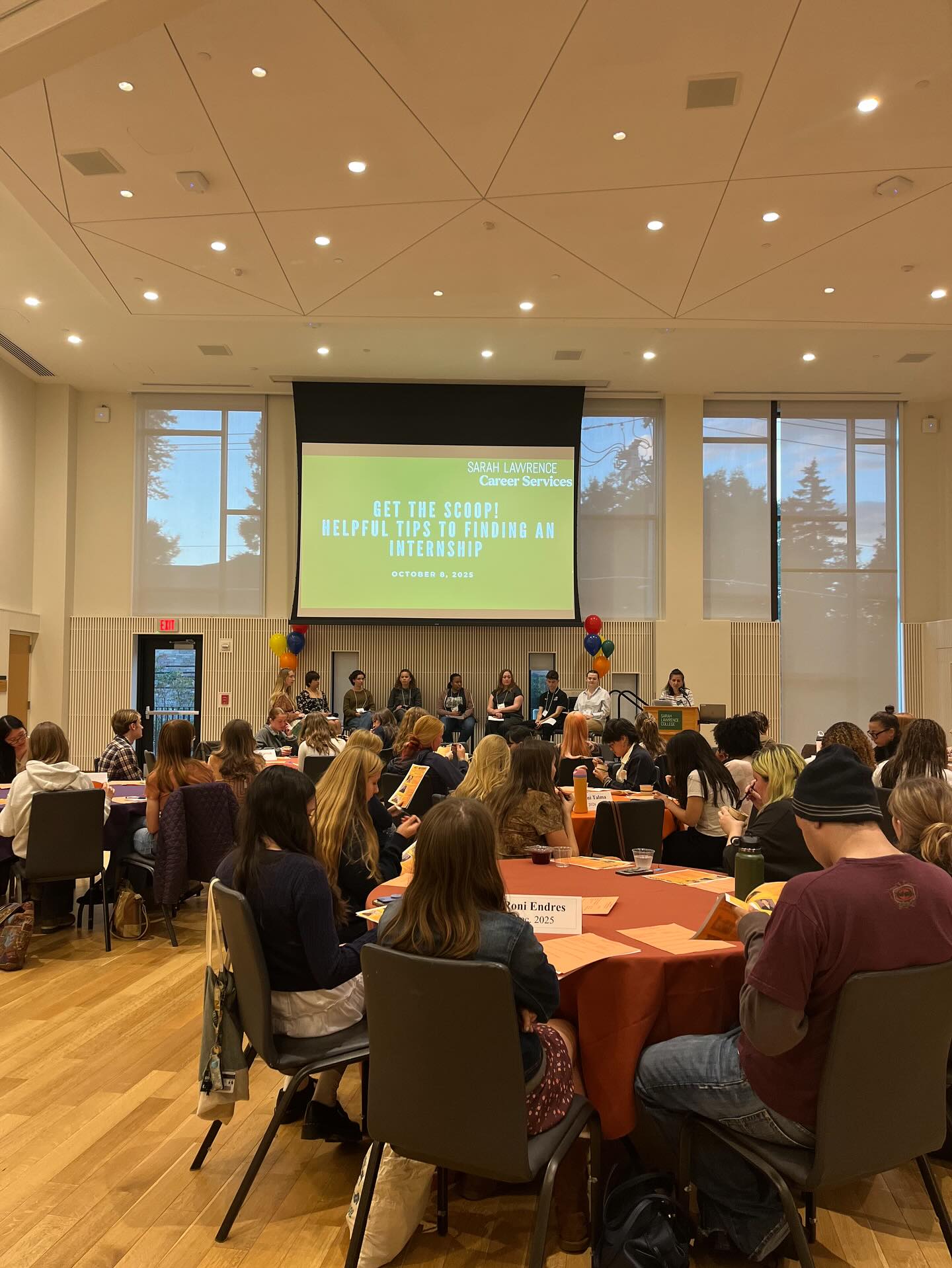 People seated at round tables in a conference room with a screen displaying "Get the Scoop! Helpful Tips to Finding an Internship, October 9, 2023."