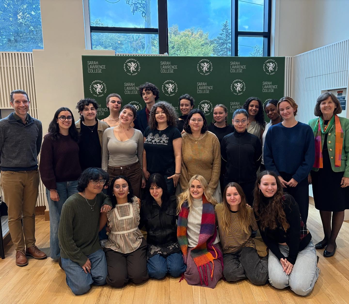 A group of people pose in front of a Sarah Lawrence College backdrop in a brightly lit room with large windows.
