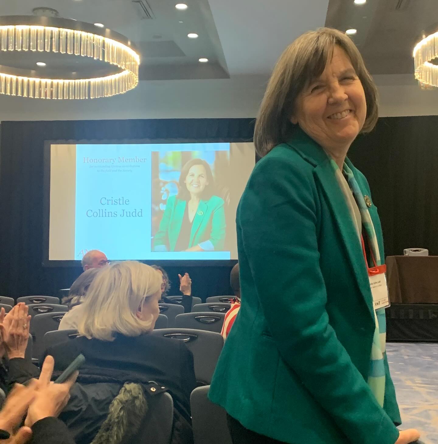 Person in a teal jacket smiles in a conference room. A screen in the background shows an honorary member's image and name. Audience claps.