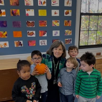 A group of children and an adult stand indoors. One child holds a pumpkin. Colorful art is displayed on the wall behind them.