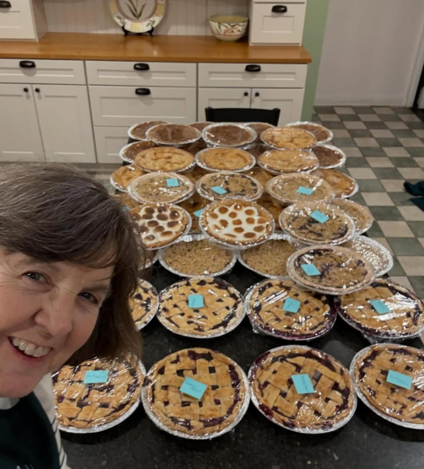 Person smiling with a table full of homemade pies, each labeled with notes, in a kitchen with white cabinets and a tiled floor.