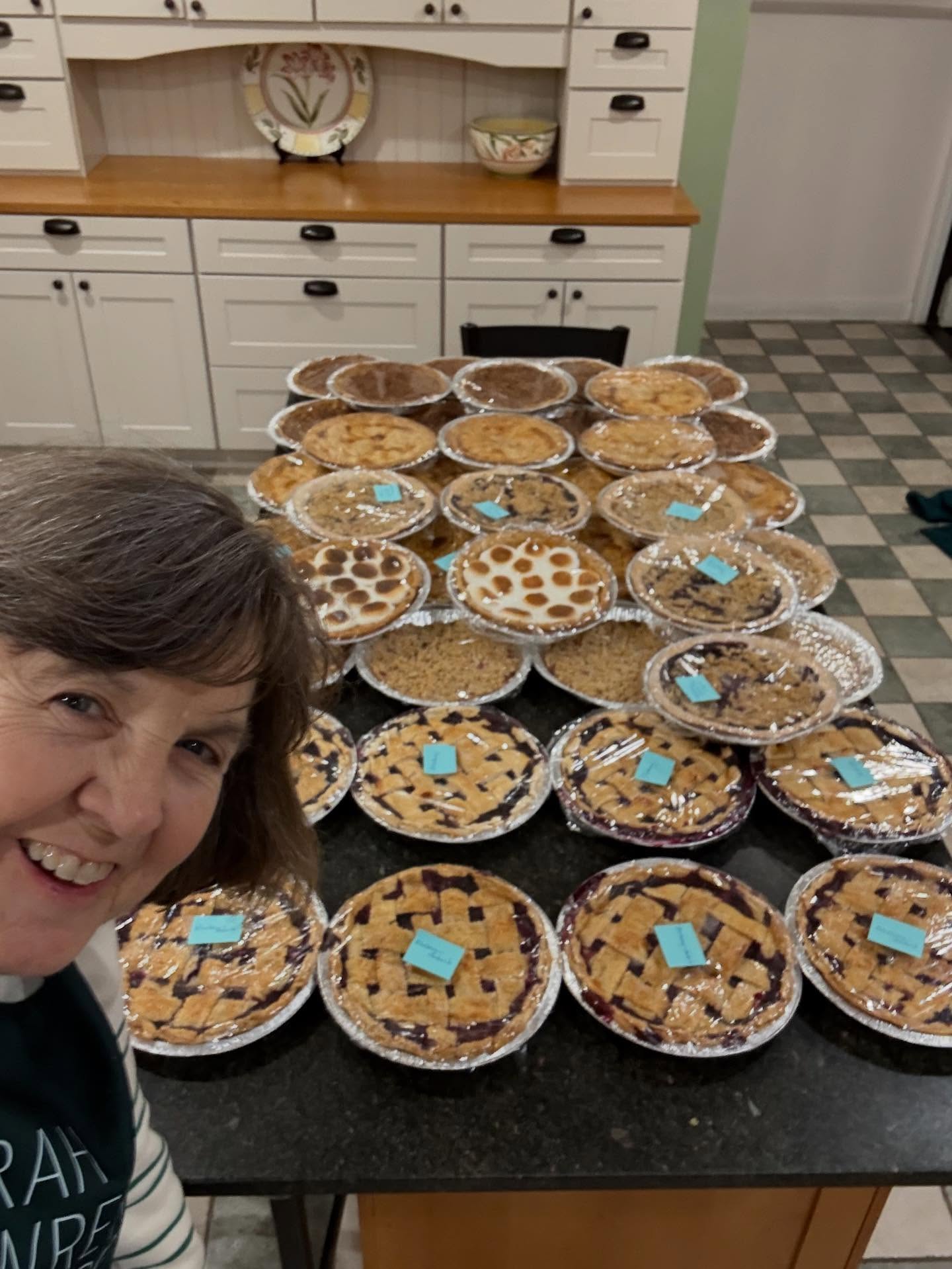 Smiling person in a kitchen with over twenty assorted pies on the counter, some with lattice tops, others with various toppings, each labeled.