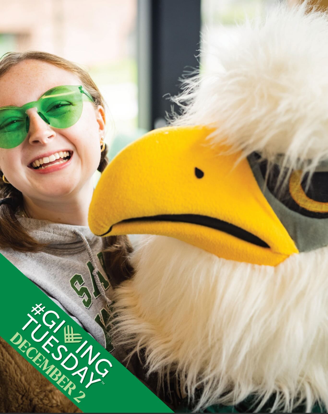 Person with green sunglasses smiles beside a large bird mascot. Text: &quot;#GivingTuesday December 2.&quot;