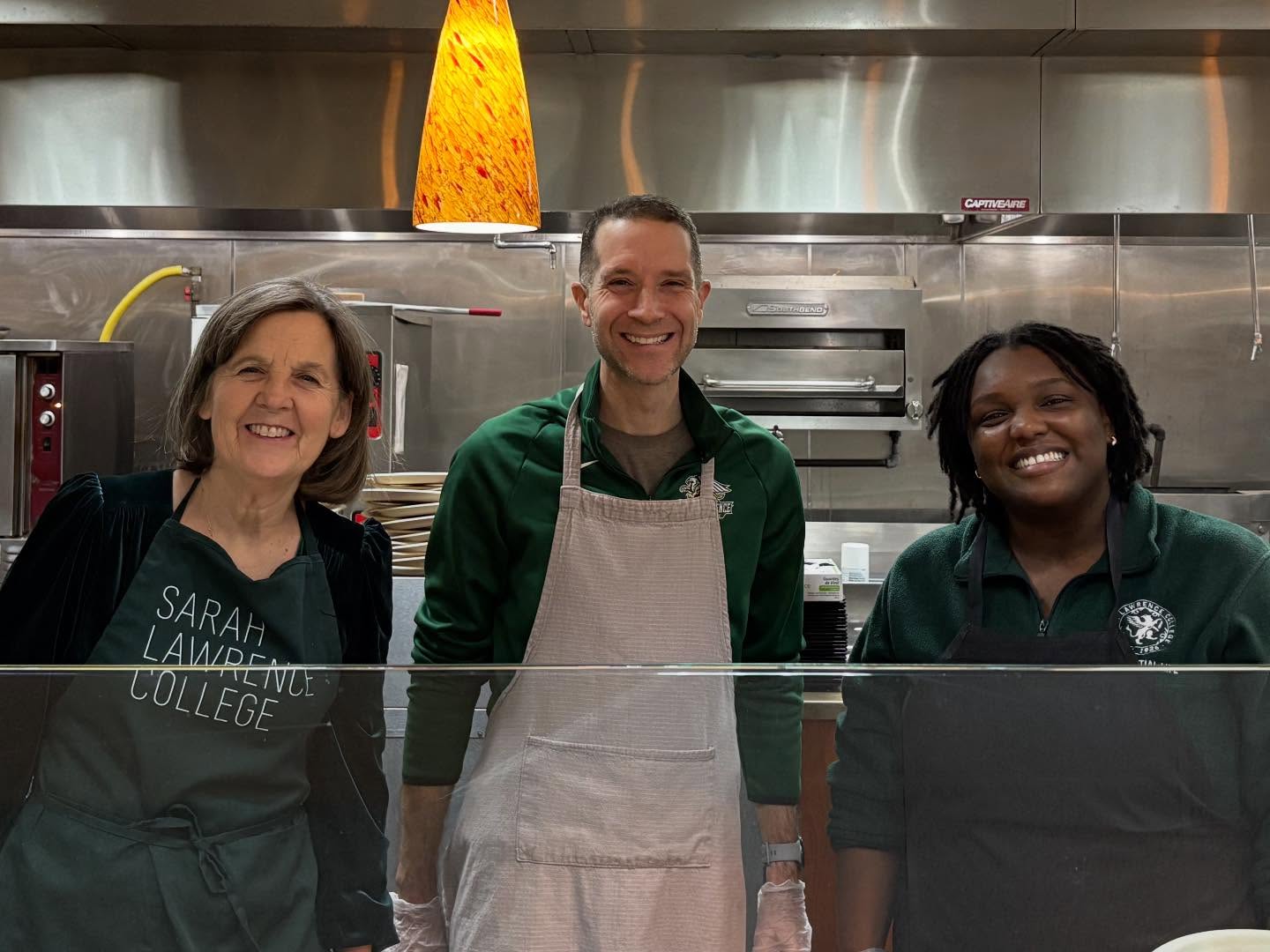 Three individuals in a kitchen wear aprons and smile at the camera. One apron reads "Sarah Lawrence College."