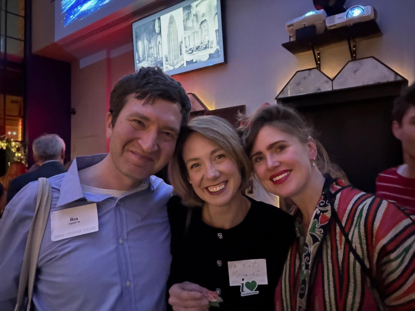 Three people smiling at an indoor event, wearing name tags. A screen and decorative lights are visible in the background.