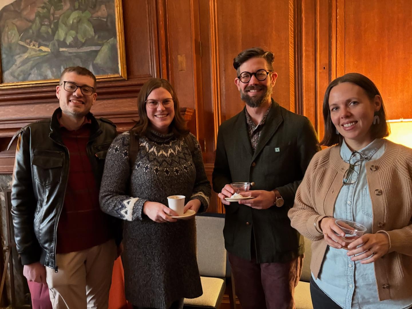 Four people smiling indoors, standing in front of a wooden wall with a painting. Two hold cups, and one has glasses hanging from a collar.